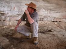 John Parkington sitting on a log, looking to the left, wearing a hat in front of old wall